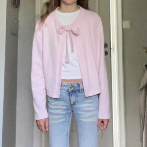 Ljusrosa kofta med rosett från 157 - Supersöt ljusrosa kofta från 157 i stickat material. Koftan har lång ärm och knyts framtill med en rosett vid halsen. Perfekt att slänga över en topp för en gullig och trendig look. Passar dig som gillar en feminin och enkel stil.