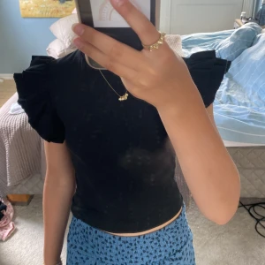 Svart volangtopp från Gina Tricot - Supersöt svart topp från Gina Tricot med ribbad struktur och volangärmar som ger en extra cool touch. Modellen är croppad och har rund hals. Perfekt för dig som vill ha något stilrent och trendigt!