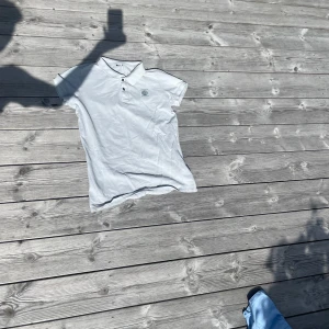 Vit pikétröja från Guess - Vit pikétröja från Guess med klassisk krage och tre knappar framtill. Tröjan har korta ärmar och en broderad logotyp på bröstet. Perfekt för en clean och stilren look. Materialet känns som bomull och är mjukt mot huden.