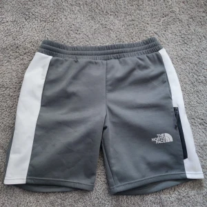 Grå-vita shorts från The North Face - Snygga grå shorts från The North Face med vita paneler på sidorna. De har elastisk midja, dragkedjeficka och sportig look. Perfekta för träning eller chill dagar. Materialet är mjukt och stretchigt för extra komfort.