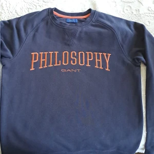 Mörkblå GANT sweatshirt Philosophy - Mörkblå sweatshirt från GANT med orange broderad text 'PHILOSOPHY' på bröstet. Klassisk rund halsringning med ribbade muddar och orange detalj på insidan av kragen. Perfekt för dig som gillar stilrena college-tröjor.