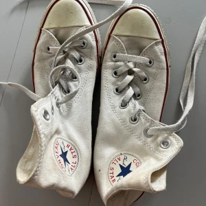 Vita Converse Chuck Taylor All Star - Klassiska vita Converse Chuck Taylor All Star high-tops med röd och blå rand längs sulan. Skorna har snörning, rund tå och är tillverkade i textil med gummisula. Perfekta för en avslappnad och cool streetstil. Storlek 38. 