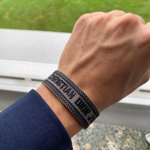 Christian Dior J'Adior armband gråbrun - Snyggt vävt armband från Christian Dior med texten 'J'Adior' i svart på grå bakgrund. Armbandet har en enkel och stilren design med justerbar knytning. Perfekt accessoar för att lyfta din outfit med en lyxig touch.