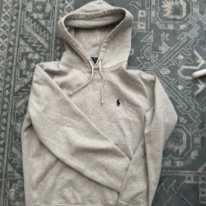 Grå hoodie från Polo Ralph Lauren XS - Snygg ljusgrå hoodie från Polo Ralph Lauren i bomull med klassisk huva och snörning. Tröjan har en broderad marinblå logga på bröstet och känguruficka framtill. Perfekt för en chill och stilren look.