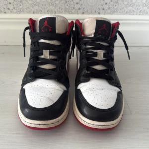 Nike Air Jordan 1 Mid Svart/Vit/Röd - Säljer ett par Nike Air Jordan 1 Mid med klassisk färgkombination i svart, vitt och rött. Skorna har svart snörning, röd swoosh och detaljer, samt vit mellansula. Ovandelen är i skinn och de har en rund tå och platt sula. Perfekta för dig som gillar streetstyle och ikoniska sneakers.