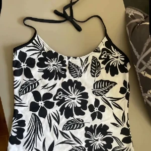 Svartvit blommig linnetopp - Snygg linnetopp med svartvita blommor och tunna axelband som knyts i nacken. Toppen har ett tropiskt blommönster och är perfekt för varma dagar. Lätt och luftig känsla, passar till både jeans och kjol.