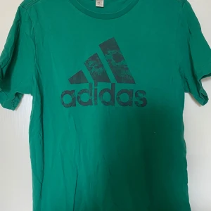 Grön Adidas t-shirt  - Snygg grön t-shirt från Adidas med stor svart logga framtill. Klassisk rund halsringning och korta ärmar. Tillverkad i mjuk bomull som känns skön mot huden. Perfekt för dig som gillar sportig och avslappnad stil. Frakt tillkommer