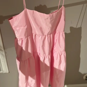 Rosa volangklänning från H&M Divided - Supersöt rosa kortklänning från H&M Divided med tunna axelband och flera volanglager. Klänningen har en luftig passform och är perfekt för varma dagar. Tillverkad i ett lätt, glansigt material som ger en fräsch och ungdomlig look.