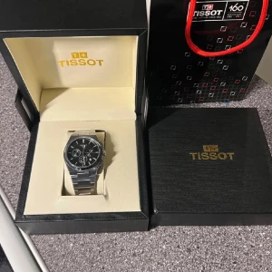 Tissot PRX kronograf i stål - Säljer en snygg Tissot PRX kronograf med svart urtavla och robust armband i rostfritt stål. Klockan har flera subdials, safirglas och klassisk länkdesign. Kommer i originalask med Tissot-logga och tillhörande påse. Perfekt accessoar för dig som gillar stilren design.