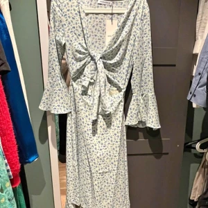 Adoore - Supersöt långklänning med blått blommönster på vit botten från Adore. Klänningen har v-ringning, knyt framtill, volangärmar och en luftig passform. Perfekt för sommarens alla tillfällen!