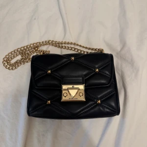 Svart quiltad axelväska Michael Kors - Snygg svart axelväska från Michael Kors med quiltad design och guldfärgade detaljer. Väskan har kedjerem och coolt lås framtill med små nitar. Perfekt storlek för det viktigaste och ger en lyxig vibe till din outfit.