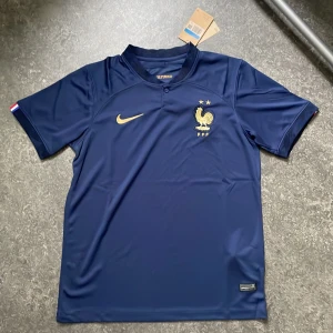 Nike Frankrike Hemmatröja VM 2022 – Storlek M - Officiell Nike Frankrike landslagströja – hemmatröjan från VM 2022 i mörkblått med exklusiva gulddetaljer. Ikoniska tupp-emblemet med två stjärnor (världsmästare!). Tröjan är i storlek M och i nytt skick- ej använd.  ✔️ Slim fit-passform ✔️ Material: 100 % polyester ✔️ Perfekt för matchdagar eller som stilren streetwear