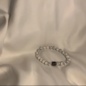 Vitt pärlarmband med marmormönster - Stilrent armband med vita pärlor och gråa marmordetaljer, samt en svart cylinderformad pärla som accent. Perfekt för dig som gillar minimalistiska accessoarer med en twist. Elastiskt band gör det enkelt att ta på och av.