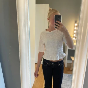 Vit smockad blus med volangkant - Supersöt vit blus med smockad midja och volangkant nertill. Modellen är croppad och har korta ärmar samt rund halsringning. Perfekt för dig som gillar en feminin och trendig look.