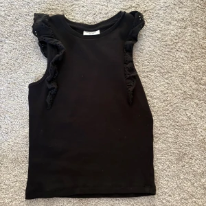 Svart ärmlös topp med volang - Svart ärmlös topp från Kappahl med snygga volangdetaljer på axlarna. Perfekt för dig som gillar en stilren look med en twist. Mjuk bomullskänsla och rund halsringning. Storlek 146/152💓