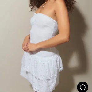 Vit axelbandslös klänning Gina Tricot - Supersöt vit kortklänning från Gina Tricot med smockad överdel och volangig kjol. Axelbandslös modell med broderade detaljer och lätt struktur i tyget. Perfekt för varma dagar när du vill känna dig extra fin. Klänningen är slutsåld på hemsidan