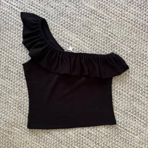 Svart oneshoulder topp med volang H&M - Säljer en svart oneshoulder topp från H&M i storlek M. Toppen har en bred volang längs den asymmetriska axeln och är figurnära. Perfekt för dig som vill sticka ut med en cool och trendig look.