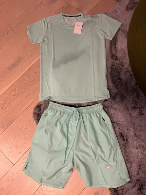 Mintgrönt träningsset från Nike - Snyggt mintgrönt träningsset från Nike med t-shirt och shorts. T-shirten har rund hals och svart Nike-logga på bröstet, shortsen har resår och dragsko i midjan samt liten Nike-logga. Perfekt för löpning eller gym. Materialet är lätt och andas. Passar M mer kroppsnära 