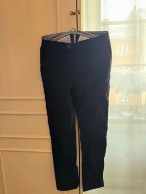 Svarta kostymbyxor John Henric - Snygga svarta kostymbyxor från John Henric med klassisk rak passform och knappgylf. Byxorna har diskreta fickor och är tillverkade i ett mjukt tyg som ger en stilren look. Perfekta för dig som vill ha en clean och modern stil.
