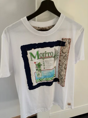 Vit t-shirt med mojito-tryck från BOB - Vit t-shirt från BOB med färgglatt mojito-tryck och dekorativa tygdetaljer framtill. T-shirten har klassisk passform och korta ärmar. Perfekt för dig som gillar unika och kreativa plagg med coola detaljer.