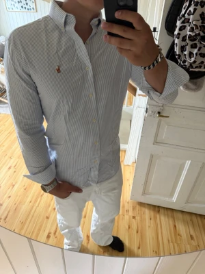 Blårandig skjorta med knappkrage - Snygg blårandig skjorta med klassisk knappkrage och broderad logga på bröstet. Skjortan har långa ärmar och vita knappar framtill. Perfekt till jeans eller chinos för en clean look.