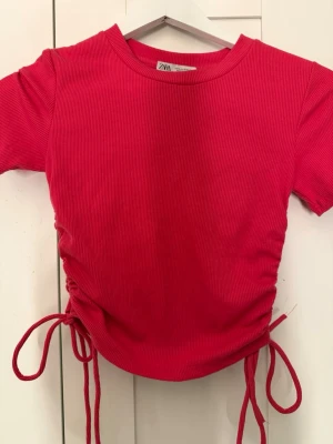 Ribbad rosa  topp med snörning från Zara - Trendig ribbad rosa  topp från Zara med korta ärmar och snörning i sidorna som ger en snygg croppad effekt. Perfekt för dig som vill ha en enkel men ändå unik topp till din outfit.