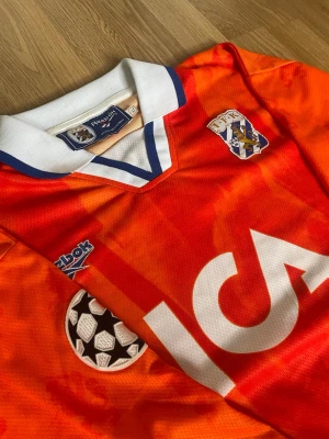 IFK Göteborg retrotröja Reebok orange - Säljer en retro långärmad fotbollströja från IFK Göteborg i orange med vita och blå detaljer. Tröjan har klubbmärke, Reebok-logga, Champions League-märke och ICA-tryck på bröstet. Klassisk krage i vitt och blått. Perfekt för samlare eller fans!