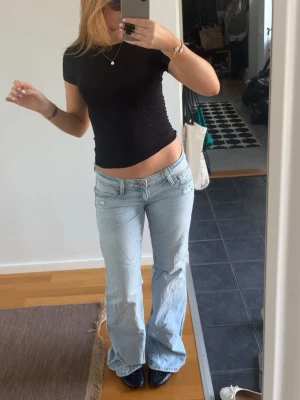 Ljusblå low waist bootcut jeans - Säljer ett par ljusblå jeans med låg midja och bootcut passform. Jeansen har klassiska fem fickor och slitna detaljer vid fickorna. Perfekta för en avslappnad och trendig look. Materialet är denim i bomull. Storlek S tall. Kan nog va lite slitna längst ner. Skriv för bild 