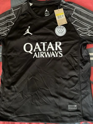 Svart PSG fotbollströja Jordan - Säljer en svart PSG fotbollströja från Jordan med vita detaljer och tryck. Tröjan har Paris Saint-Germain-logga, Qatar Airways sponsortryck och coola mönster på ärmarna. Tillverkad i lätt och ventilerande Dri-FIT-material, perfekt för träning eller match.