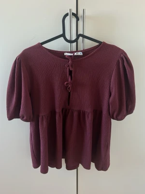 Vinröd blus med puffärm och knyt - Säljer en vinröd blus med puffiga korta ärmar och rund halsringning. Blusen har knytning framtill och ett lätt rynkat fall nedtill. Tillverkad i ett mjukt, strukturerat tyg som ger en avslappnad men ändå söt look.