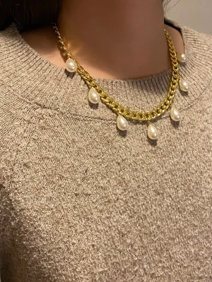Guldhalsband med droppformade pärlor - Säljer ett chunky halsband i guldfärgad rostfritt med droppformade vita pärlor som hänger längs hela kedjan. Halsbandet har en modern och trendig look som sticker ut och passar perfekt till lager-på-lager-stil.