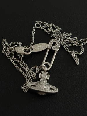 Silverhalsband - Snyggt halsband i silverfärg från med ett ikoniska orb-hänget dekorerat med små gnistrande stenar. Kedjan är tunn och har ett unikt säkerhetsnål-inspirerat fäste. Perfekt för dig som gillar statement-smycken och vill sticka ut.