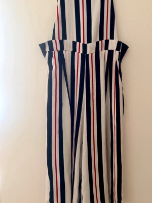 Vit jumpsuit med ränder från Stradivarius - Snygg jumpsuit med vertikala ränder i svart, vitt och rött. Modellen har vida ben och markerad midja med bred linning. Tillverkad i ett lätt och luftigt tyg som känns skönt mot huden. Perfekt för dig som gillar en trendig och avslappnad stil.