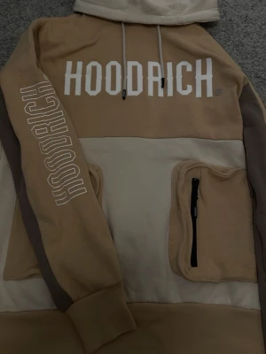 Hoodrich hoodie - Snygg hoodie i beige och creme från Hoodrich med stor logga på bröstet och ryggen. Säljer denna för att ja köpte den för 4 år sedan på Jd sports så den passar mig inte längre 