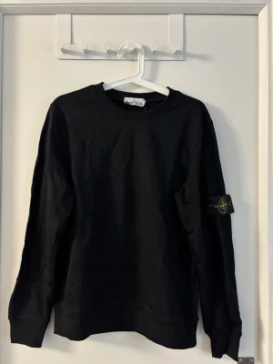 Svart sweatshirt från Stone Island - Säljer en svart sweatshirt från Stone Island med den ikoniska loggan på vänster ärm. Tröjan har rund halsringning, ribbade muddar och är tillverkad i mjukt bomullsmaterial. Perfekt för en clean och stilren look.