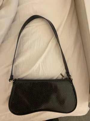 Svart axelväska  - Snygg svart axelväska med ormmönstrad yta och silverfärgad dragkedja. Väskan har en smal axelrem och en stilren, avlång form som ger en trendig look. Perfekt för dig som gillar minimalistisk och edgy stil.