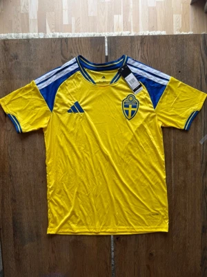 Sverige matchtröja Adidas gul/blå - Gul matchtröja från Adidas med blå detaljer och Sveriges landslagsemblem på bröstet. Tröjan har rund hals, blå ränder på axlarna och är tillverkad i ett lätt, tekniskt material som andas. Perfekt för fotboll eller träning.