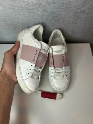 Valentino opens  - Snygga vita sneakers från Valentino med ett brett ljusrosa band över vristen och klassiska nitar på sulan. Skorna har rund tå, vita snören och är tillverkade i läder. Perfekta för dig som vill ha en stilren men ändå unik look. Mycket fint skick storlek 35.5 passar 36/36.5