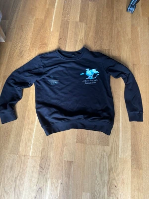 Svart sweatshirt med tryck och brodyr - Svart sweatshirt med rund hals och ribbade muddar. På bröstet finns ett stort tryck med blå moln, grå bokstäver och texten '16 Janvier 2020, 14h30 Jardin des Tuileries'. Perfekt för en chill och trendig look.