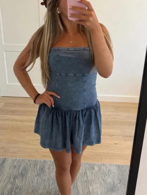 Blå axelbandslös jeansklänning - Säljer en söt axelbandslös kort jeansklänning i blått med markerad midja och volang nedtill. Klänningen har en enkel, stilren design och är perfekt för varma dagar. Passformen är normal och materialet är jeans vilket ger en cool och avslappnad look.