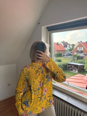 Gul blommig blus med hög krage - Snygg gul blus med färgglatt blommönster i rosa, blått och grönt. Blusen har hög krage och långa ärmar, perfekt för dig som vill sticka ut med en färgklick i garderoben. Tillverkad i ett lätt och mjukt tyg som känns skönt mot huden.finns två knappar man knäpper bak vid nacken. Tillkommer en extra knapp i tvättrådslappen. 