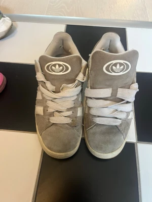 Adidas campus - Säljer ett par gråa Adidas high top sneakers i mocka med vita detaljer och breda vita skosnören. Skorna har den klassiska Adidas-loggan på plösen och tre vita ränder på sidan. Perfekta för dig som gillar retrostil och streetwear.