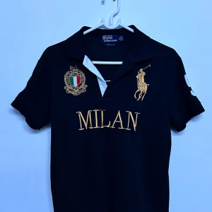 Chief keef Svart piké t-shirt från Polo Ralph Lauren med MILAN-tryck -  Vintage chiefkeef polo Svart piké t-shirt från Polo Ralph Lauren med guldigt MILAN-tryck och broderad logga på bröstet. Nummer 5 på ryggen och klassisk krage med knappar. Perfekt för dig som gillar sportig stil. 