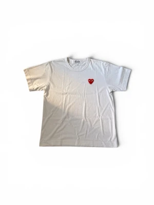 Vit t-shirt Comme des Garçons Play - Säljer en klassisk vit t-shirt från Comme des Garçons Play med det ikoniska röda hjärtat med ögon broderat på bröstet. T-shirten har rund hals och normal passform. Perfekt för dig som gillar stilrena plagg med en twist. De står m på tröjan men de är för att de är m i barn storlek. De finns små fläckar i mitten av tröjan