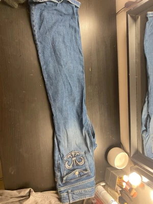 Blå jeans med broderad bakficka - Snygga blå jeans med en unik broderad detalj på bakfickan och knappstängning. Klassisk femficksmodell med raka ben och bälteshällor. Jeansen har en cool tvättad look och passar perfekt till en avslappnad stil.