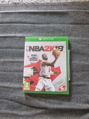 NBA 2K18 till Xbox One - Säljer spelet NBA 2K18 till Xbox One. Omslaget är vitt och rött med en basketspelare i action på framsidan. Spelet är i fysisk utgåva med grön plastbox och PEGI 3-märkning. Perfekt för dig som gillar basket och sportspel.