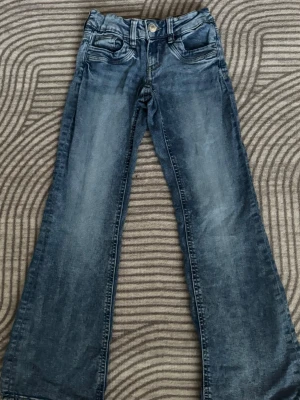 Blå bootcut jeans - Ett par blå bootcut jeans med klassisk femficksmodell. Jeansen har normal midja och vida ben, vilket ger en avslappnad och trendig look. Tillverkade i denim med snygg tvättad effekt och knappgylf.