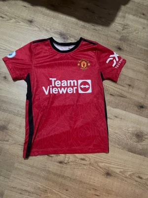 Manchester United matchtröja Casemiro - Röd Manchester United matchtröja med svarta detaljer och tryck av TeamViewer på bröstet. Nummer 18 och Casemiro på ryggen. Korta ärmar, rund hals och klubbmärke på vänster bröst. Tillverkad i lätt och ventilerande material, perfekt för fotbollsträning.