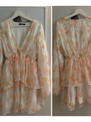 Blommig kortklänning med volang - Söt kortklänning med blommigt mönster i ljusa toner av rosa, gul och grön på vit botten. Klänningen har v-ringning, långa ärmar och volangdetaljer. Tillverkad i ett tunt, lätt material som ger en luftig känsla.