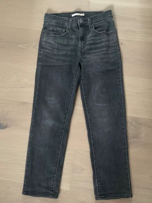 Levi 724 High Rise Straight - Snygga svarta jeans med raka ben och klassisk femficksmodell. Jeansen har subtila slitningar och en mörk tvätt som ger en cool look. Tillverkade i ett mjukt denimtyg som sitter skönt hela dagen. Storlek W26 